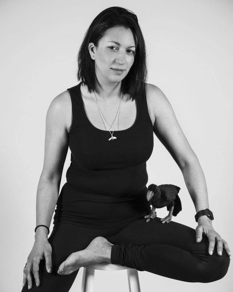 A woman sitting on a stool wearing all black. She has dark straight hair and a crow on her lap