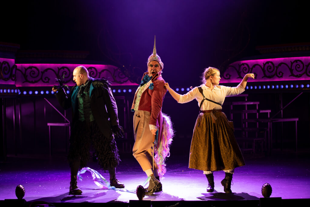 Three actors are together posing as strong people. Hamish the unicorn is in the middle and is singing.