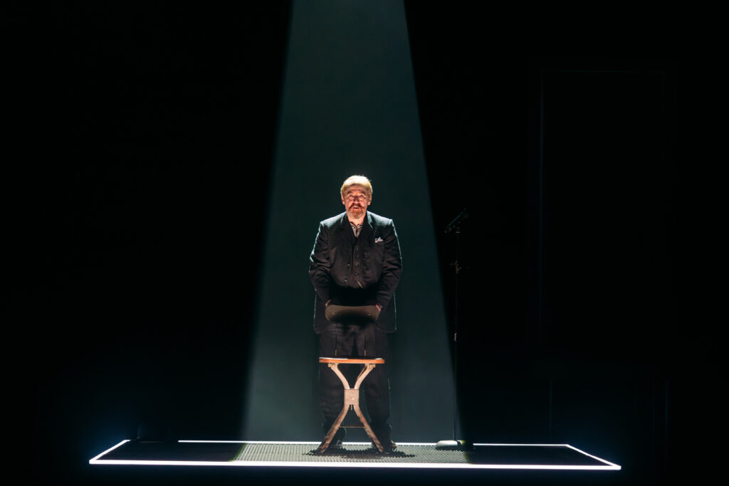 Actor Forbes Masson stands in a shaft of light. The rest of the stage is dark.