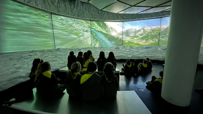 A group of children are watching a set of screens displaying information about icebergs.