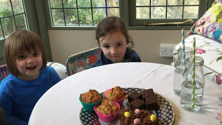 Two three-year-old children sit looking at a plate of cakes. They are in a garden house.