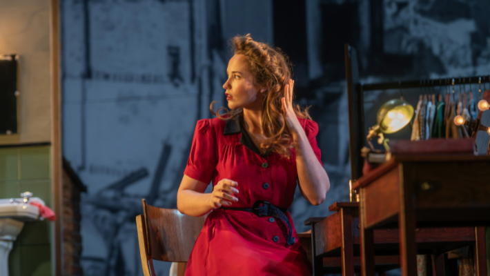 A woman sits looking to the left of the image. She wears a red dress and is fixing her hair. She is acting in a play at the Lyceum in Edinburgh. The Girls of Slender Means.