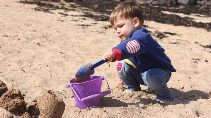 A toddler playing with a bucket and spade on a sandy beach in Scotland.