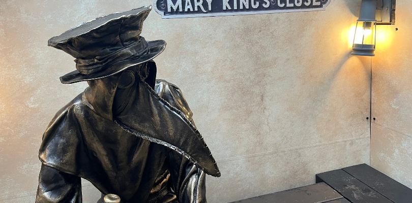 A statue of a plague doctor is sat on a bench. The plague doctor has a long beak mask and a top hat.