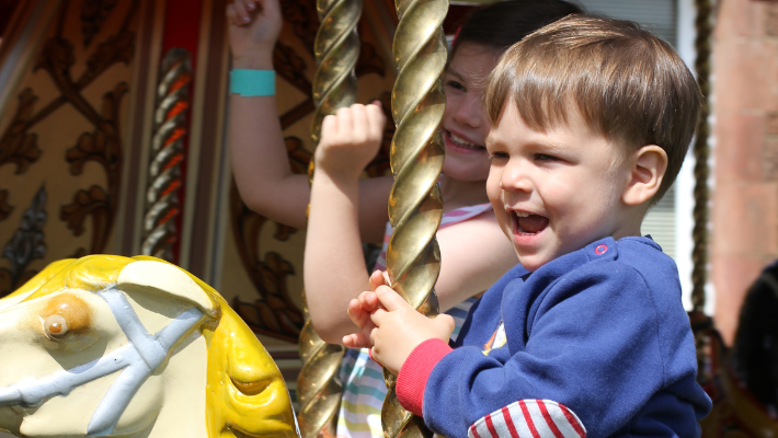 A toddler smiling on a carrousel ride. He is wearing a blue jumper.