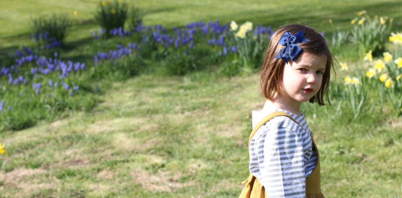 A 5 year old girl stands looking at the camera side on. Behind her are daffodils and bluebells.