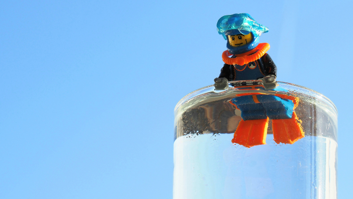 A LEGO figure dressed as a diver is propped up on the edge of a glass of water.