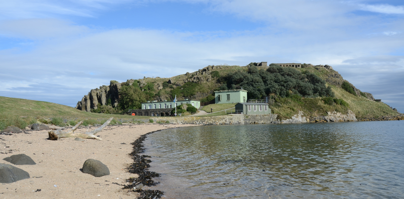The view from the beach on inchcolm island. The waves lap the shore with some buildings in the background.