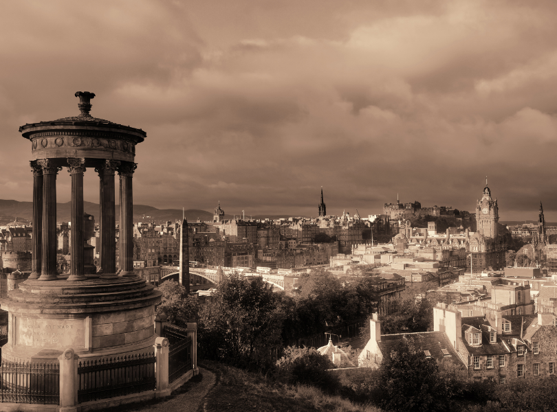 Sepia-toned view of Edinburgh from Calton Hill, featuring the Dugald Stewart Monument in the foreground and a panoramic cityscape with Edinburgh Castle and church spires in the distance.