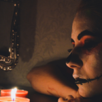 Person with dark Halloween face paint rests near glowing red candles, their face illuminated by soft candlelight, creating a spooky and eerie atmosphere.